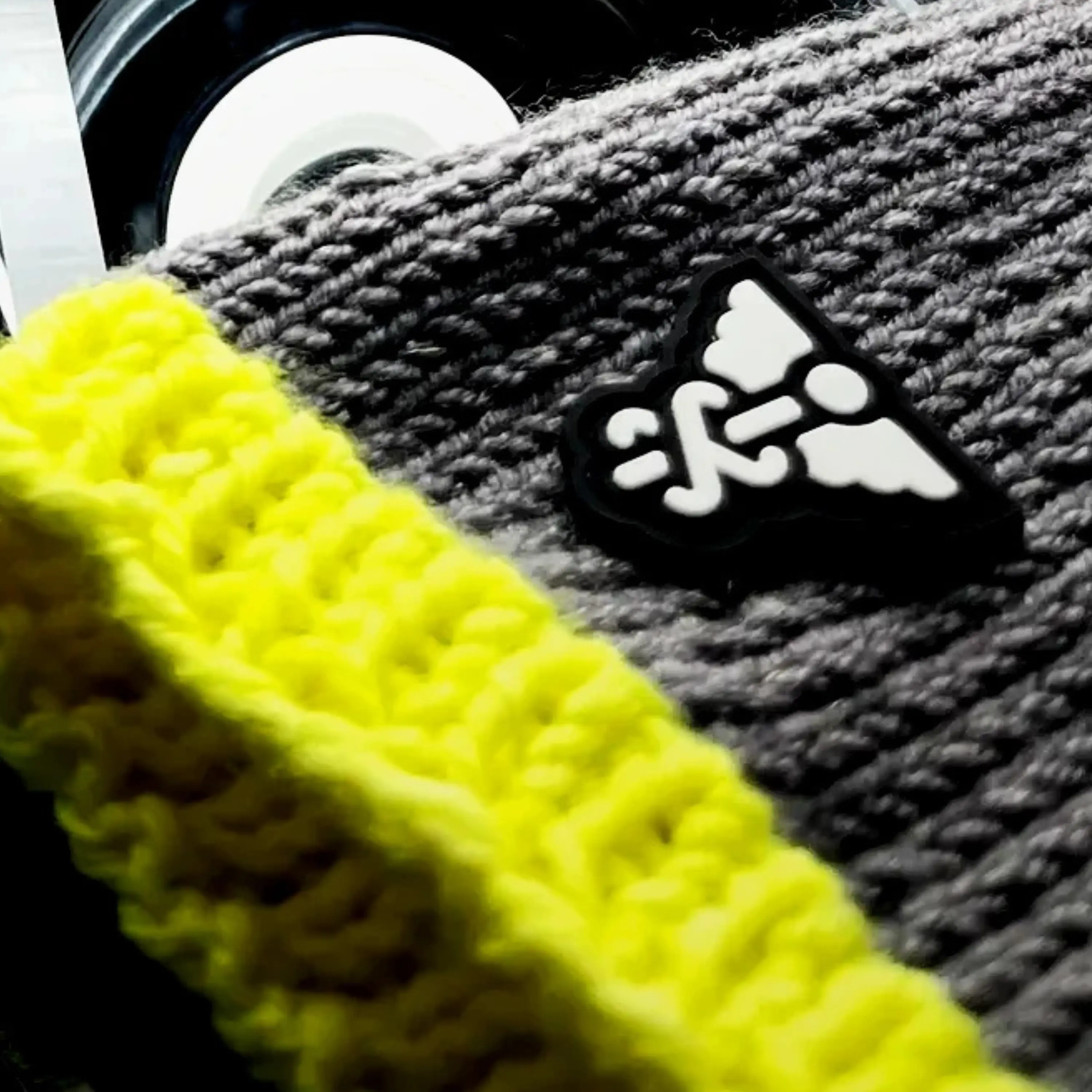 Close-up of THE CILICE: grey knit with the symbol patch sewn flat, a neon yellow-green knit band out of focus in front.